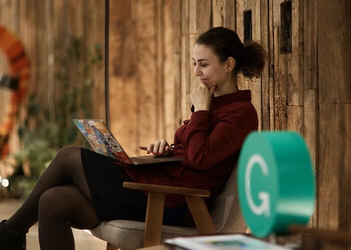 A person in a collared shirt, skirt, and tights sits in a cushioned chair smiling down at an open laptop resting on their lap. The walls have wood paneling and soft light illuminates plants in the background. A 3D Grammarly logo sits on a table in the foreground.