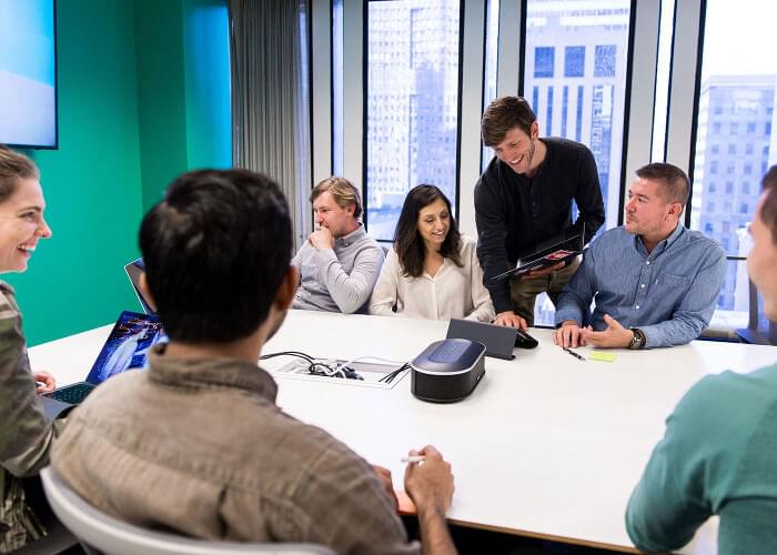 Seven people sit around a conference table smiling at each other while some work on laptops. City views can be seen outside a large window in the background.